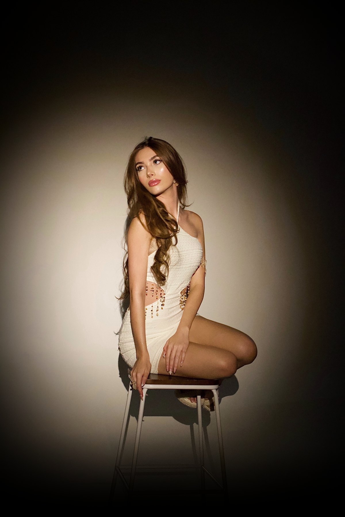 Daniella in a white beaded dress, seated on a stool under a spotlight
